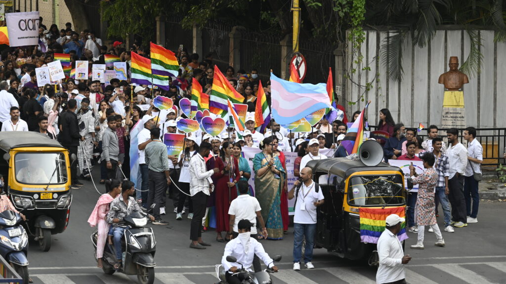 People participating in pride walk, waving pride flag & trans flag, led by an autorickshaw with a speaker