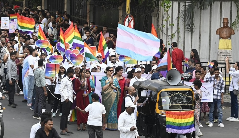 People participating in pride walk, waving pride flag & trans flag, led by an autorickshaw with a speaker