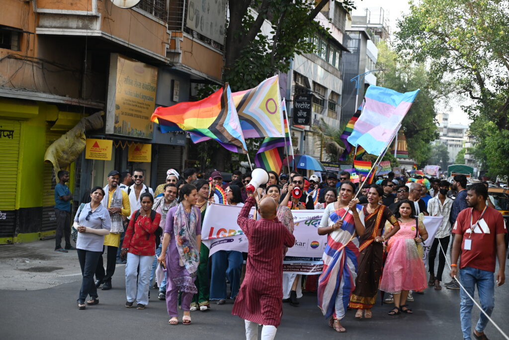People marching cheerfully in a pride walk, waving rainbow flag, transgender flag, and intersex-inclusive progress pride flag.