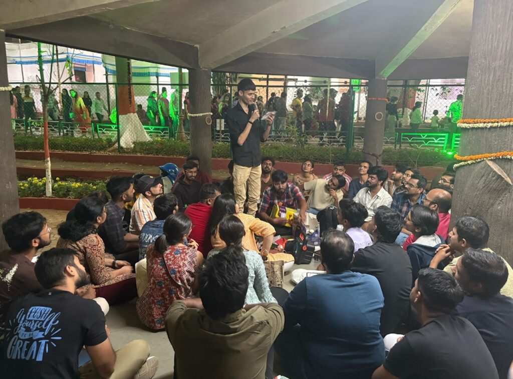Group of people sitting in a circle. They are listening to a person standing in the centre reading from a note.